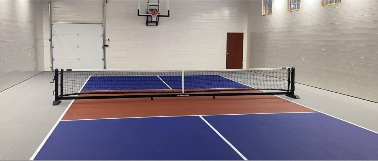 An indoor pickleball court in a white court with basketball hoops overhead. 