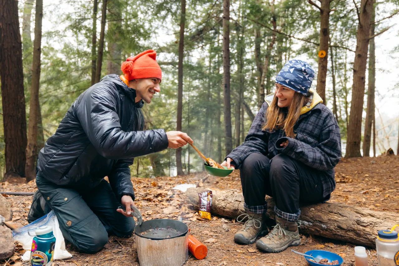 A man and woman cook food on top of a campfire at a campsite during midday.