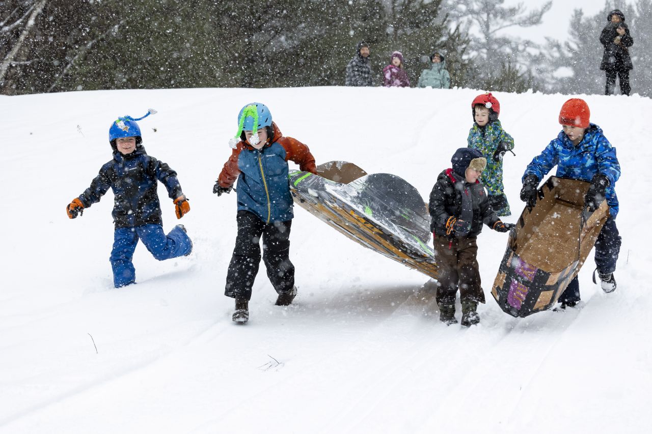 Four energetic young boys in brightly colored winter gear run down a snowy hill, carrying two homemade cardboard sleds towards the start of a cardboard sled race.
