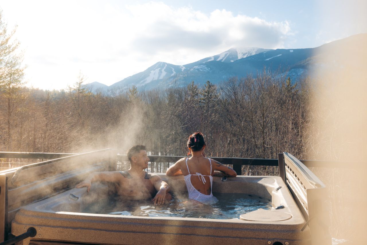 Man and woman in bathing suits sit in hot tub with champagne flutes on winter day. Woman's back is turned as she looks out at mountain views and the man looks at her.