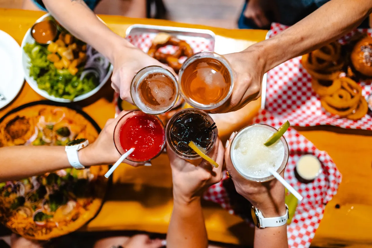 A close-up view looking straight down at five hands clinking drinks glasses together over a restaurant table of food.