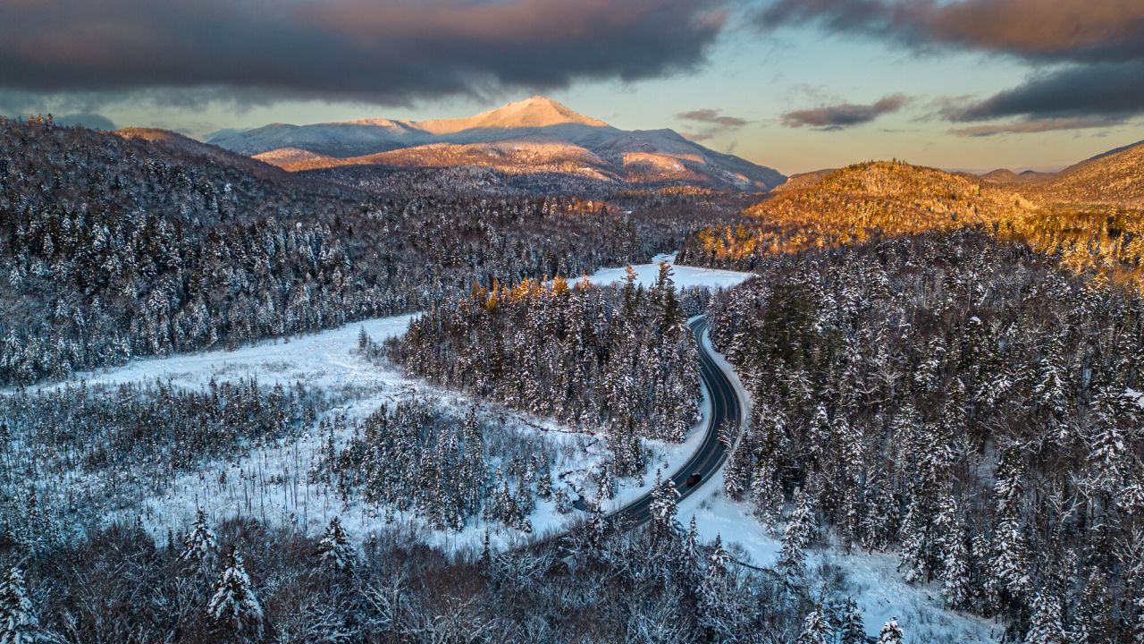 Aerial view at sunrise or sunset of snowcapped Whiteface Mountain in the distance with road, forests, and marshland covered in snow in the foreground