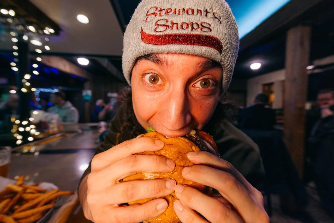 A young man wearing a Stewart's Shops beanie hat makes a startled face while biting into a cheeseburger.