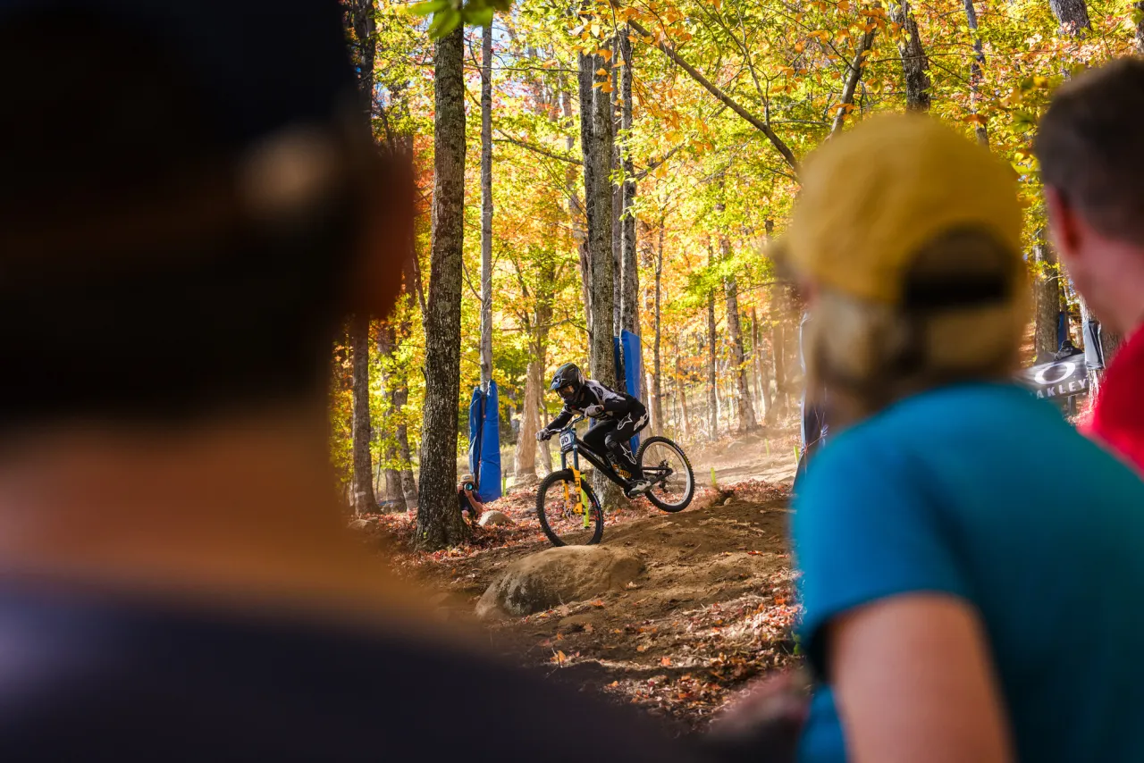 Mountain biker competing on trail through woods with spectators looking on