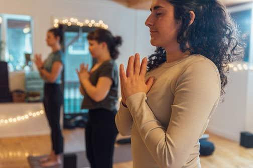 Three people practice yoga in a calm studio, standing with palms together in relaxed focus. Soft lighting and string lights create a peaceful ambiance.