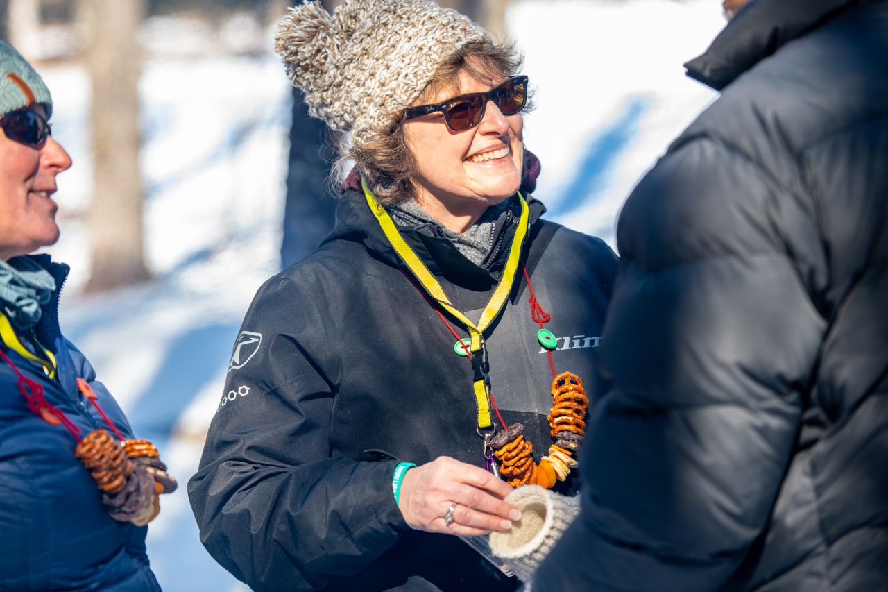 A woman in winter outdoor clothing and wearing a necklace made of pretzels smiles in the woods on a sunny winter day.