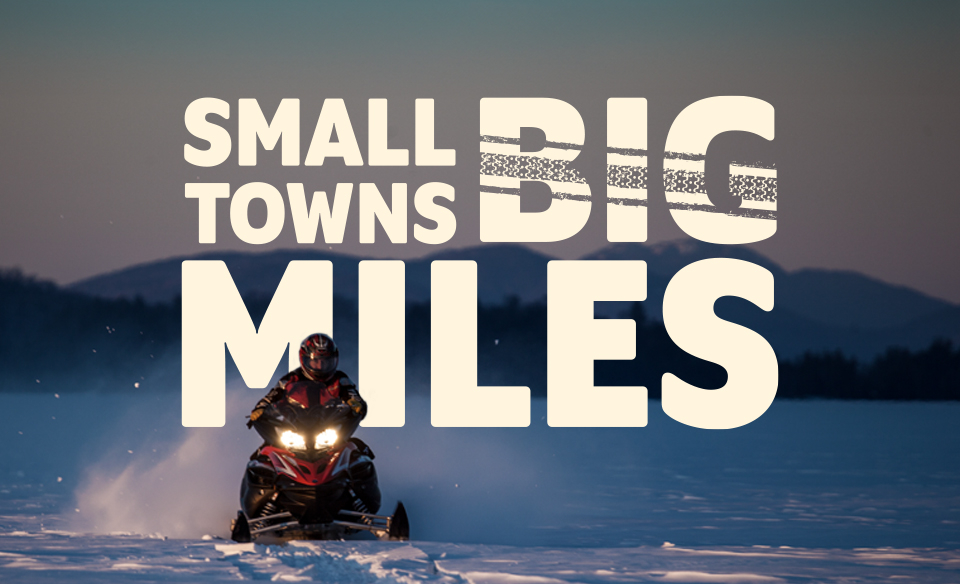 A graphic with the words: Small towns big miles, is edited behind a snowmobiler on an icy lake. 