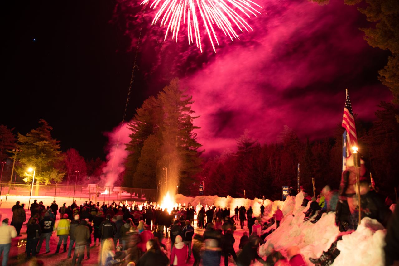 Vibrant red fireworks explode over a large crowd gathered around a bonfire at night.
