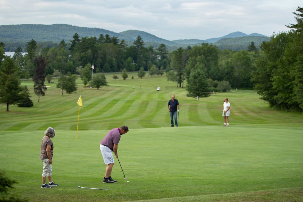 A group of people golf on a green course with mountains in the background. 