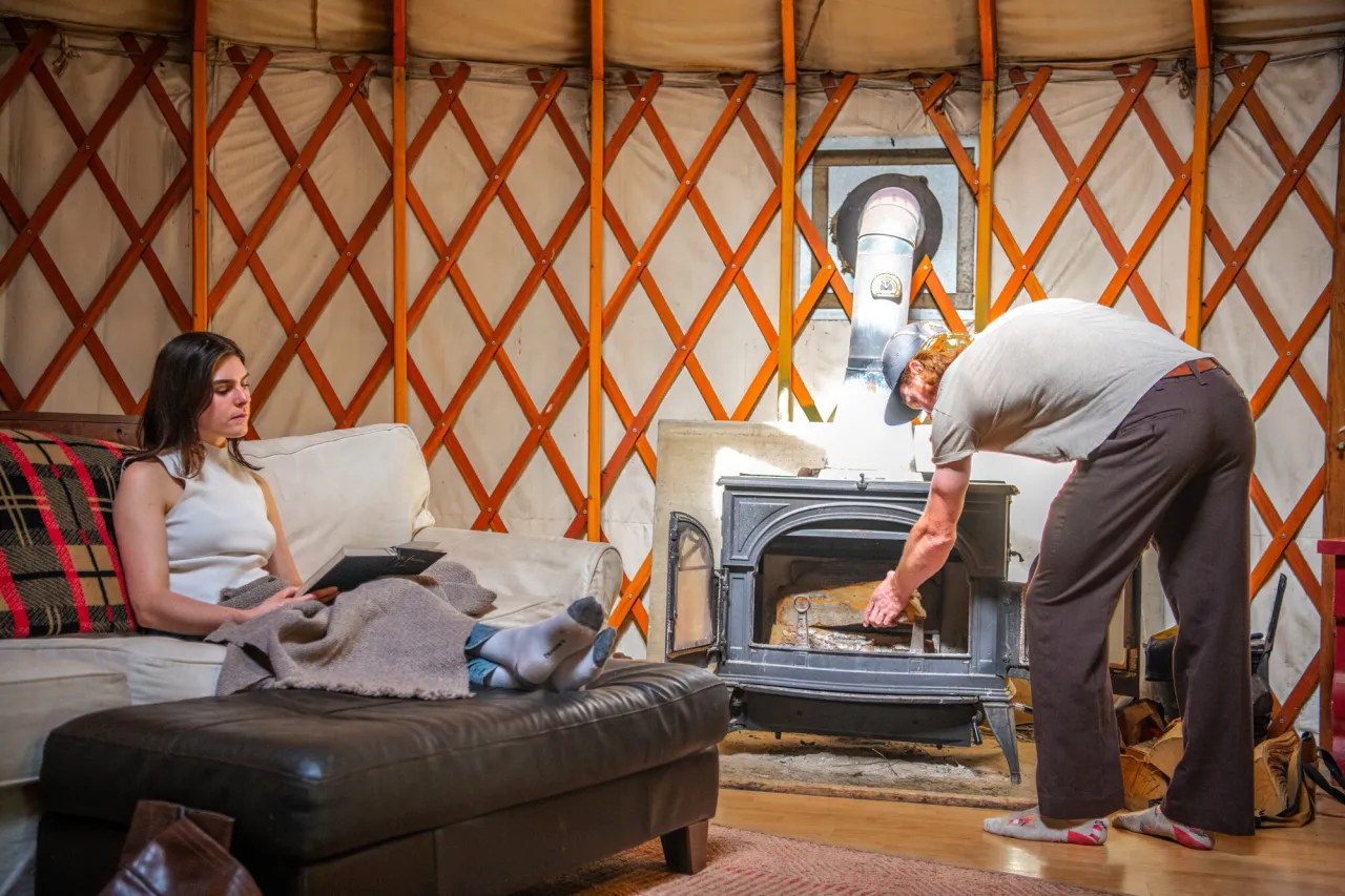 A man loads a fireplace in a cabin while a woman sits on a sofa.