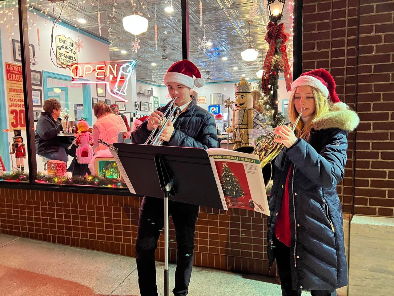 Two teenagers wearing Santa hats play musical instruments outside a brightly-lit retro diner.