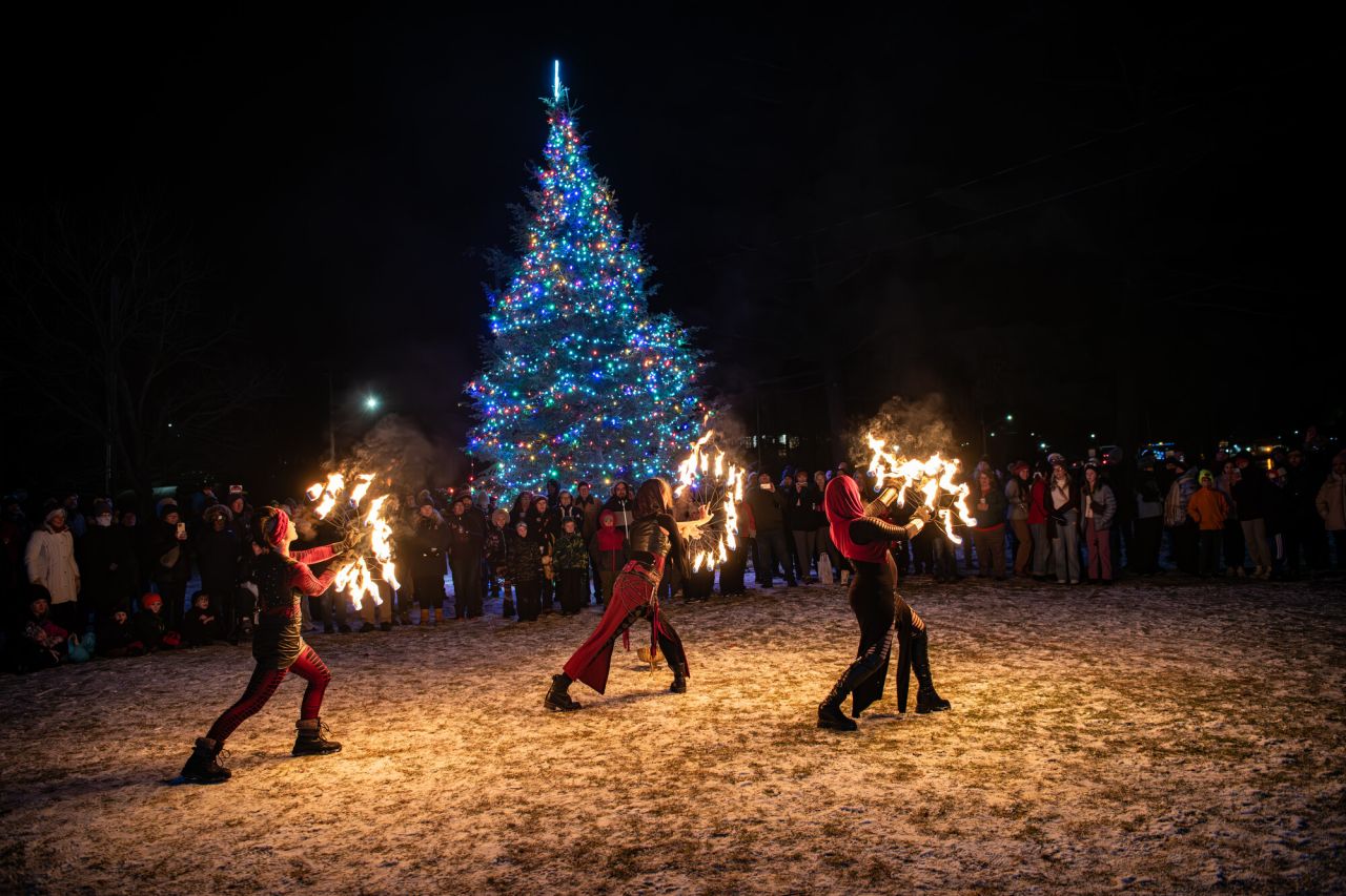 Three fire dancers perform for a crowd in front of a large lit up Christmas tree in winter. 