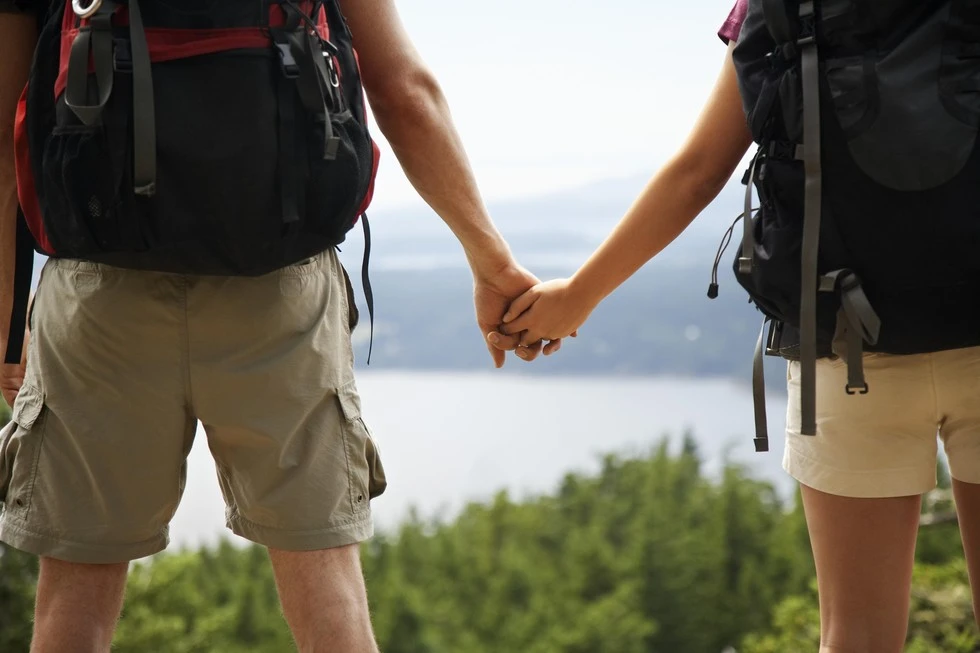 Two people hold hands with backpacks on and looking at a view with trees and a body of water.