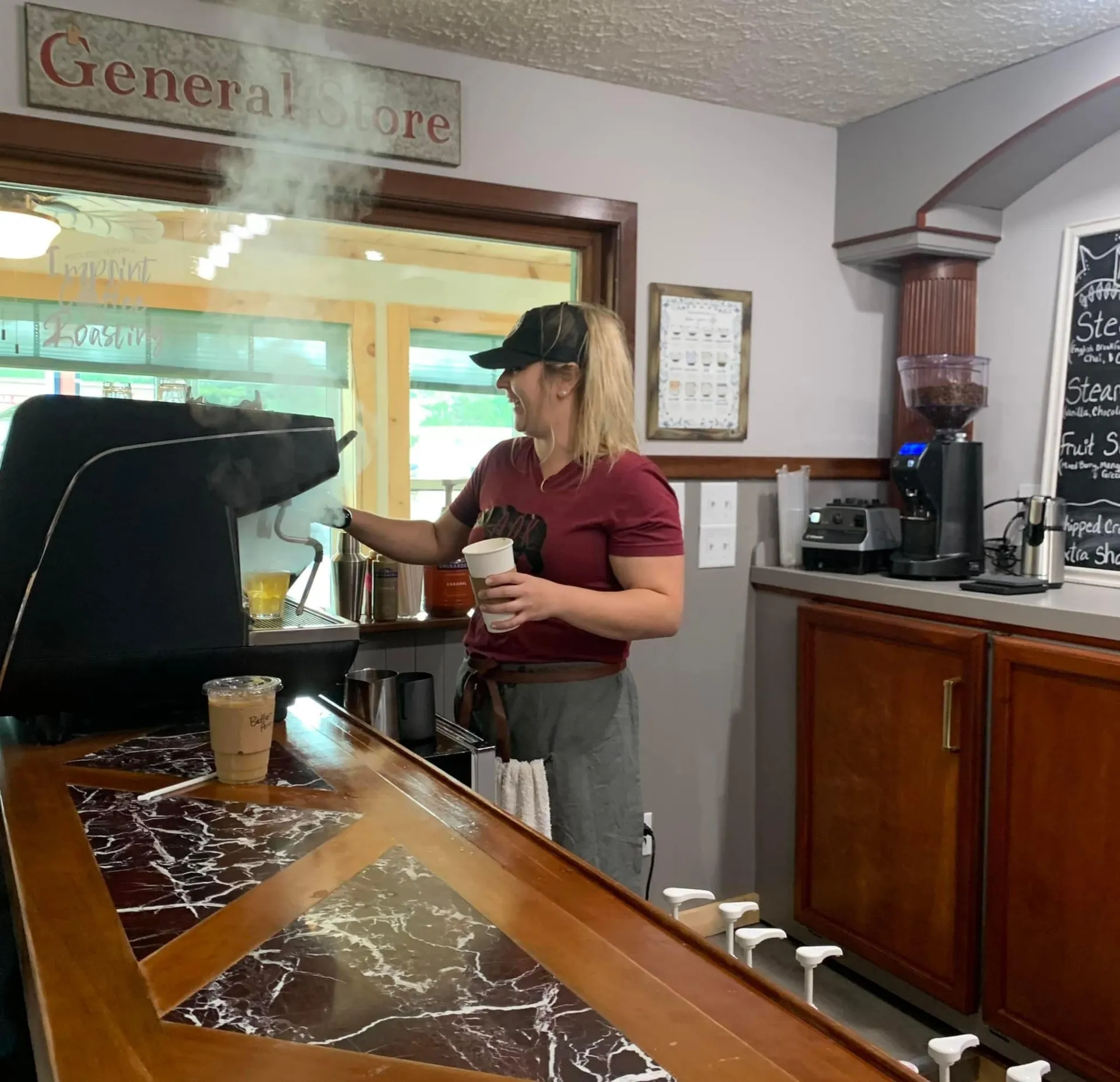 A happy blond woman operates the espresso machine while steam rises from the preparation.