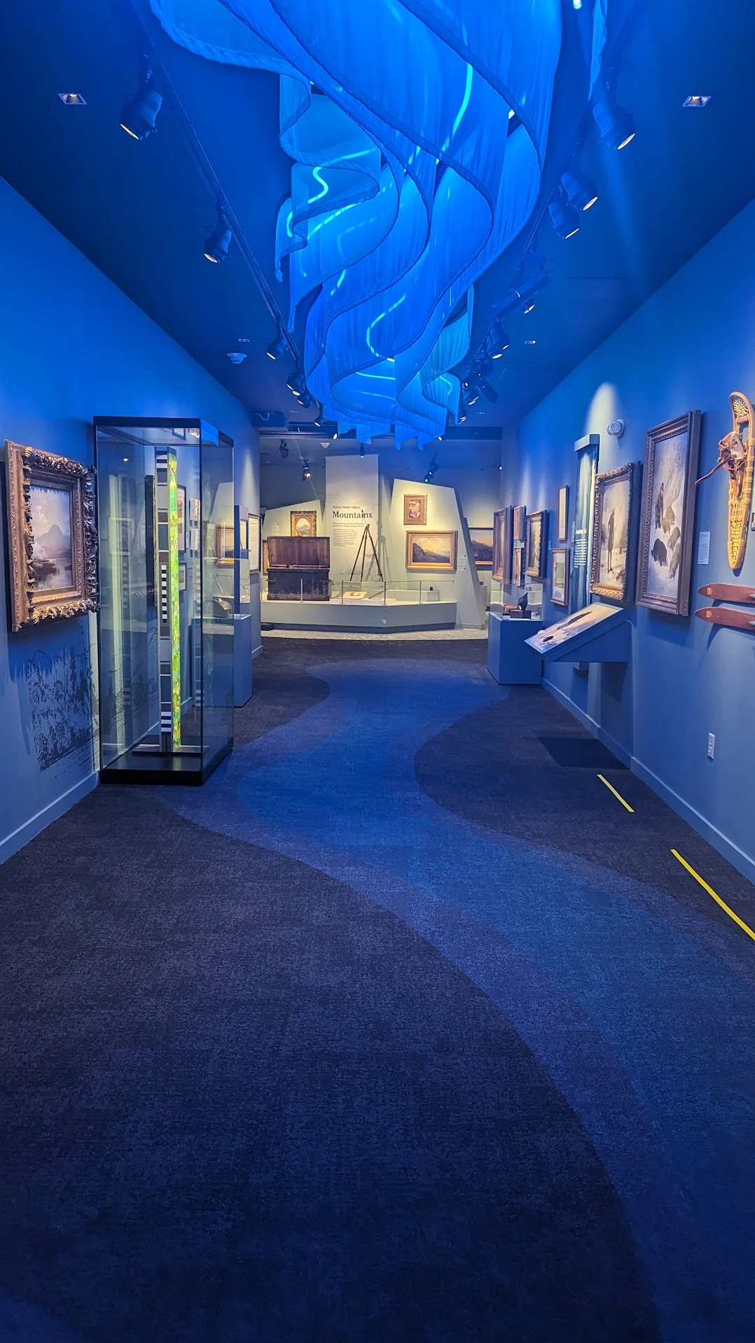 Art pieces on display in a long hallway with blue walls, a blue carpet with a winding pathway design and blue light fixture that resembles waves