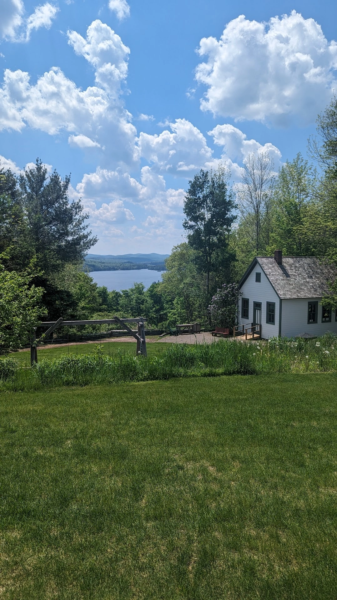 Blue Mountain Lake under billowing white clouds across a lush green lawn in front of a quaint white cabin.