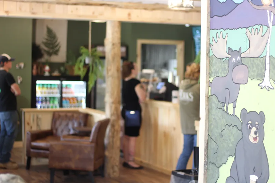 A view of the mural on the wall depicting a moose, bear and other wilderness scene with a set of leather chairs in a cozy corner, the drink fridge and people lined up to get their order in.