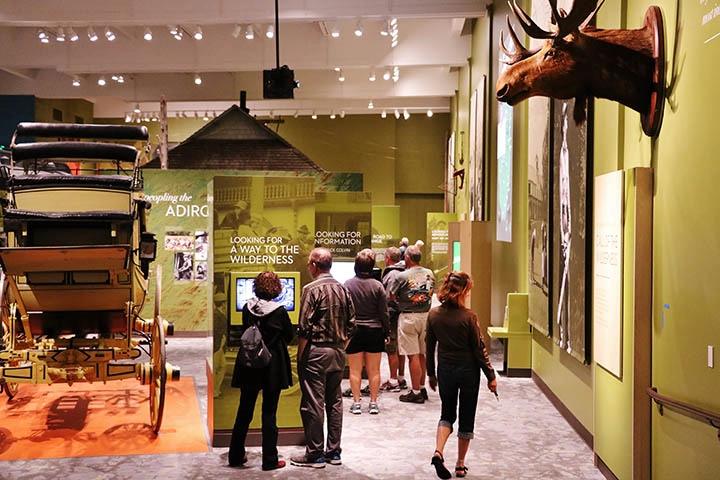 A large room with green walls and high ceilings where people are viewing the plaques under various displays like a carriage and a moose.