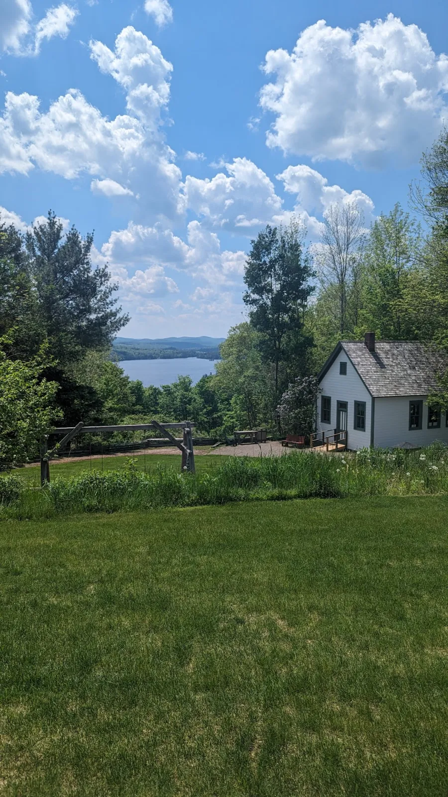 Blue Mountain Lake under billowing white clouds across a lush green lawn in front of a quaint white cabin.
