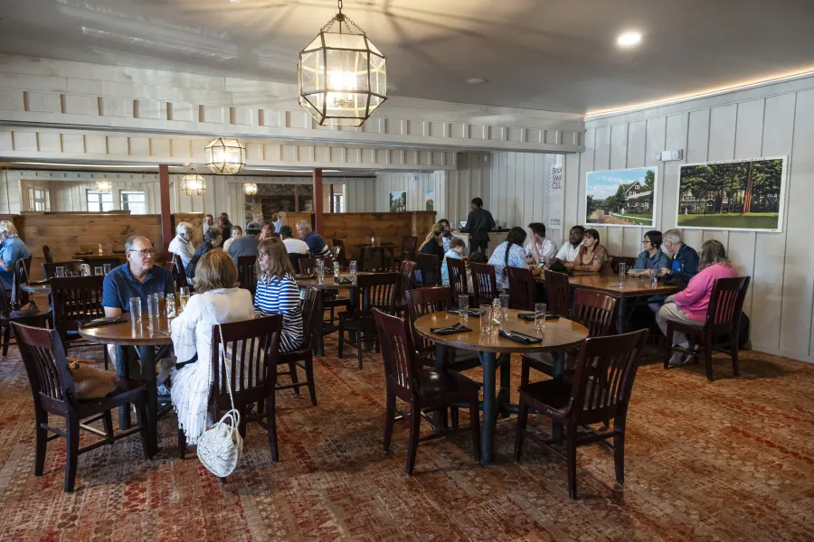 A grand dining room with simple chandeliers and white paneled walls filled with large wooden dining tables.